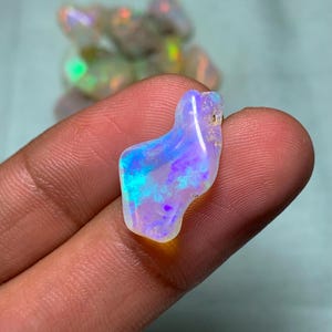 Könnte beinhalten: Ein weißer Opal-Edelstein mit blauen und violetten irisierenden Blitzen. Der Opal ist unregelmäßig geformt und hat eine glatte, polierte Oberfläche.