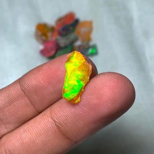 May include: A small, green and orange opal gemstone with a rough, irregular shape. The opal is being held by a finger.