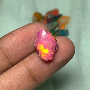 May include: A pink, yellow, and orange rough opal gemstone with iridescent flashes. The opal is held in a hand.