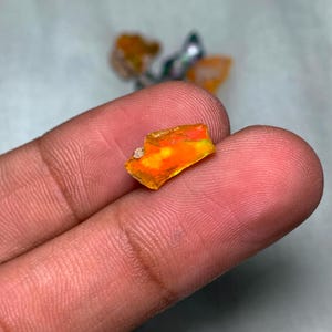 May include: A small, rough, orange and yellow opal gemstone with iridescent flashes of color. The opal is being held in a hand.