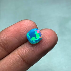 Puede incluir: Una piedra preciosa de ópalo de corte cuadrado con una vibrante exhibición de colores. La gema presenta una base azul con destellos de verde y rosa. La piedra se sostiene entre dos dedos sobre un fondo azul claro.
