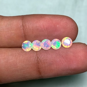 May include: Five round opals with an iridescent sheen are shown. The opals display a mix of colours, including white, pink, and green. The gemstones are presented against a neutral background.