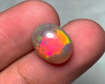 Könnte beinhalten: Ein ovaler Opal-Edelstein. Der Opal zeigt ein Farbspiel mit leuchtenden Farbtönen wie Rosa, Orange und Grün auf einem durchscheinenden Hintergrund. Der polierte Edelstein wird zwischen Fingern gehalten und zeigt seine schillernde Oberfläche.