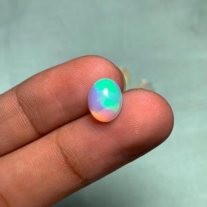 May include: An oval-shaped opal gemstone with a vibrant play of color. The opal displays a mix of green, blue, purple, and orange hues. The gemstone is held between fingers, showcasing its polished surface and iridescent qualities.
