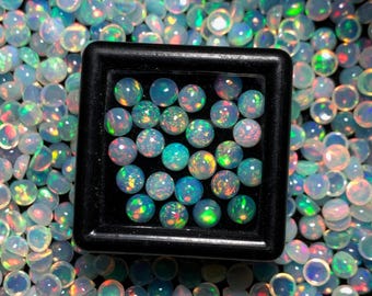 May include: A collection of iridescent opal gemstones, many with a round shape. A black square container holds a selection of the opals, showcasing their vibrant play of colour. The opals display a range of colours, including blue, green, and red.