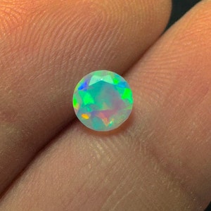 Puede incluir: Una piedra preciosa de ópalo redonda e iridiscente con un juego de colores verde claro, azul y rosa. El ópalo está engastado en un engaste redondo transparente.