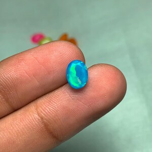 May include: An oval blue opal gemstone with a vibrant play of colour. The gemstone displays shades of turquoise and green, with a polished, faceted surface. The opal is held between fingers, showcasing its size and brilliance.
