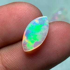K&ouml;nnte beinhalten: Ein marquise-geschliffener Opal-Edelstein mit einem durchscheinenden K&ouml;rper und lebhaften Blitzen in Neongr&uuml;n, Blau und Orange. Der Stein weist eine glatte, polierte Oberfl&auml;che und eine spitz zulaufende ovale Form auf, die das Licht reflektiert.