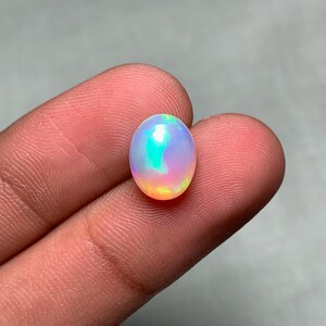 Puede incluir: Una gema de ópalo ovalada y vibrante. El ópalo muestra un juego de colores con tonos azules, verdes y naranjas. La gema se sostiene entre dos dedos sobre un fondo gris neutro.