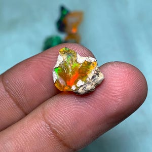 May include: A small, rough, orange and green opal gemstone with a white matrix. The opal is being held by a finger.