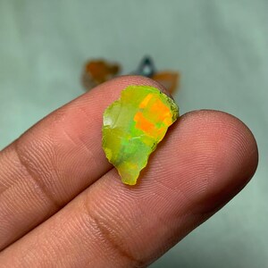 May include: A small, irregular piece of green opal with orange and yellow flashes. The opal is being held by a finger.