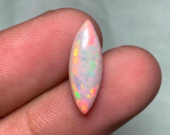 Könnte beinhalten: Ein polierter, oval geformter Opal-Edelstein. Der Opal hat eine milchig-weiße Grundfarbe mit rosa, grünen und gelben irisierenden Blitzen. Der Edelstein wird zwischen den Fingern gehalten und zeigt sein lebendiges Farbspiel.