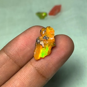 May include: A rough, orange and green opal gemstone with a black and white matrix. The opal is a raw, unpolished stone.