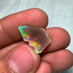 May include: A vibrant, iridescent opal gemstone fragment. The stone displays a spectrum of colors, including green, red, and yellow, with a translucent appearance. The opal is held in a hand, showcasing its natural, irregular shape.
