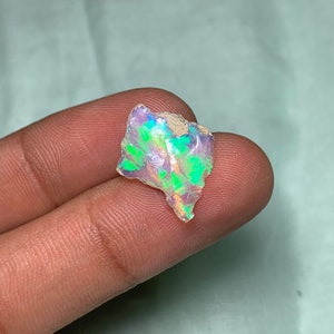 Könnte beinhalten: Ein kleiner, irisierender Opal mit grün-violettem Schimmer. Der Opal ist roh und hat eine natürliche, unregelmäßige Form.