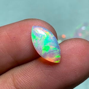 K&ouml;nnte beinhalten: Ein marquise-geschliffener Opal-Edelstein mit einem durchscheinenden K&ouml;rper und lebhaften Blitzen in Neongr&uuml;n, Orange und Gelb. Der Stein weist eine glatte, polierte Oberfl&auml;che und eine spitz zulaufende, l&auml;ngliche Form auf, die das Licht einf&auml;ngt.
