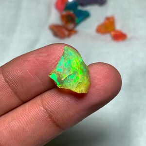 May include: A vibrant, rough-cut opal gemstone with a striking display of green and yellow hues. The stone is held in a hand, showcasing its natural, irregular shape and internal color play. Other colorful gemstones are visible in the background.
