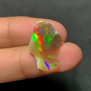 Peut inclure: Une pierre d'opale brute avec des couleurs internes vibrantes de rouge, vert et jaune. La pierre a un aspect translucide, avec un jeu de couleurs visible sur sa surface. L'opale est tenue dans une main sur un fond sombre.