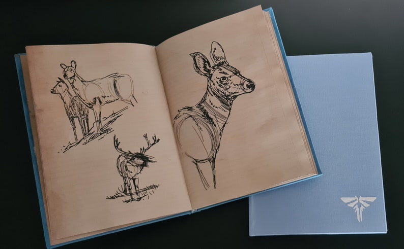 May include: An open book with hand-drawn sketches of deer on the pages. The book has a blue cover and is lying on a black surface.