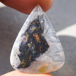 Peut inclure: Une pierre précieuse en forme de goutte d'eau avec une base blanche laiteuse translucide. La pierre présente un motif sombre et organique avec des inclusions noires, dorées et brun rougeâtre. La surface polie de la pierre est lisse et réfléchissante.
