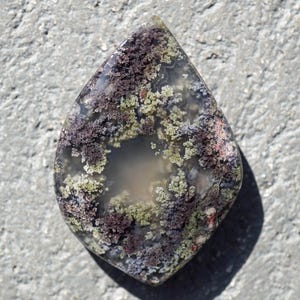 Pode incluir: Uma pedra preciosa em forma de gota com uma base translúcida, apresentando inclusões roxas escuras, verdes e brancas. A pedra polida está sobre um fundo cinza texturizado. Esta pedra única é ideal para a fabricação de joias.
