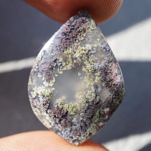 May include: A teardrop-shaped gemstone with a clear center and a surface covered in a mix of colors, including purple, green, and white. The stone is held between two fingers, showcasing its unique pattern and texture. This gemstone could be used for jewelry.