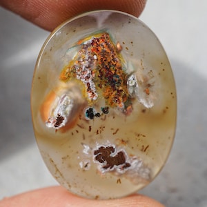 May include: Oval-shaped gemstone with a translucent, light brown base. The stone features a central inclusion of orange, white, and brown mineral deposits. Small, dark brown spots and patches are scattered throughout the stone.