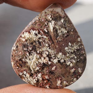 May include: A teardrop-shaped gemstone with a unique pattern. The stone features a mix of colors, including brown, white, and red, with a speckled and textured appearance. The overall effect is organic and natural, suggesting a polished stone.