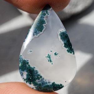 Könnte beinhalten: Ein tropfenförmiger Edelstein mit weißer Basis und komplizierten Mustern aus dunkelgrünen, moosartigen Einschlüssen. Die polierte Oberfläche reflektiert das Licht und unterstreicht die natürliche Schönheit des Steins. Der Edelstein wird zwischen zwei Fingern gehalten.