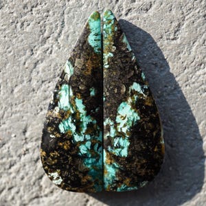 Puede incluir: Dos piedras preciosas a juego en forma de lágrima. Cada piedra presenta una llamativa combinación de colores negro, marrón y turquesa. Las piedras preciosas tienen un aspecto natural y texturizado, con una superficie pulida. Las piedras se muestran sobre una superficie gris texturizada.