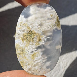 Puede incluir: Gema ovalada con apariencia nublada y en capas. La piedra presenta tonos blancos, grises y beige, que recuerdan un paisaje con formaciones tenues. La superficie pulida refleja la luz, resaltando los patrones naturales.