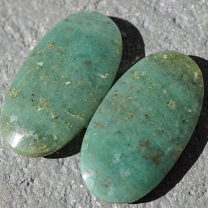May include: Two oval-shaped, polished gemstones in shades of teal and green. The stones have a smooth, glossy surface with subtle variations in color and texture. The gemstones are displayed on a gray, textured surface.
