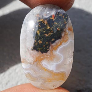 Puede incluir: Gema ovalada con una base blanca lechosa translúcida. La piedra presenta un patrón central verde oscuro y negro con acentos naranjas y rojos. Los bordes tienen un diseño en capas y ondulado con tonos naranjas y blancos. La piedra se sostiene entre dos dedos.