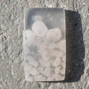 Könnte beinhalten: Ein rechteckiger, durchscheinender Cabochon-Stein mit einer glatten, polierten Oberfläche und weißen, wolkenartigen blütenförmigen Einschlüssen. Der Stein zeigt ein sanftes, milchiges Aussehen mit variierender Opazität in seiner klaren Struktur.