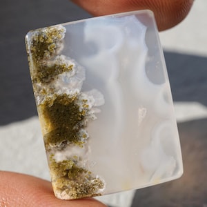 Könnte beinhalten: Ein rechteckiger, polierter Edelstein mit einer durchscheinenden weißen Basis. Der Stein weist ein auffälliges Muster aus moosgrünen und goldenen Einschlüssen entlang einer Kante auf, wodurch ein natürliches, organisches Design entsteht. Der Stein wird zwischen zwei Fingern gehalten.