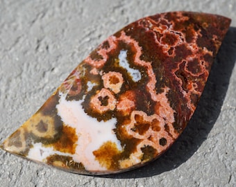 May include: A polished, teardrop-shaped gemstone with a unique pattern. The stone features a mix of colours, including white, brown, orange, and red. The surface has a marbled appearance with circular and linear patterns.