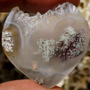 Può includere: Una pietra d'agata lucida a forma di cuore con una base traslucida bianco latte e intricate inclusioni dendritiche simili a muschio nelle tonalità del verde salvia e del viola scuro. Il bordo superiore presenta una superficie cristallina frastagliata.