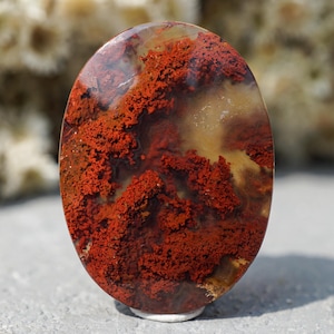 May include: An oval-shaped polished gemstone featuring a translucent, pale yellow base with deep, vibrant red moss-like inclusions spreading across the surface. The stone has a smooth, glossy finish that reflects light, highlighting the intricate, branching patterns of the red mineral deposits embedded within the clear, light-coloured quartz structure.