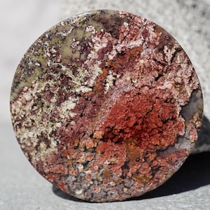 Könnte beinhalten: Ein runder, polierter Stein mit einem komplexen Muster aus roten, weißen und grünen Farbtönen. Die Oberfläche des Steins hat ein strukturiertes Aussehen mit einer Mischung aus Farben und Mustern. Der Stein ist wahrscheinlich ein Dekorationsartikel oder ein Schmuckstück.