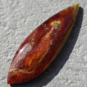 May include: A polished, elongated gemstone with a pointed end. The stone features a blend of red, brown, and yellow hues, with a glossy finish. The natural stone is approximately 10 cm long, and is displayed on a gray surface.