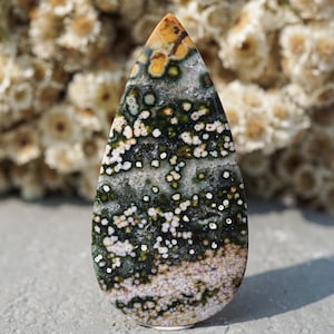 May include: A teardrop-shaped ocean jasper gemstone featuring a polished surface with a complex pattern of dark green, white, and tan circular orbs and mottled inclusions throughout the stone, tapering to a rounded point at the top.