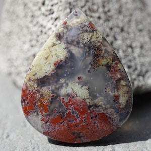 Könnte beinhalten: Ein tropfenförmiger, polierter Edelstein. Der Stein zeigt eine Mischung aus roten, cremefarbenen und grauen Farbtönen mit komplexen Mustern. Dieser Edelstein ist wahrscheinlich für Schmuck oder Dekorationszwecke bestimmt.