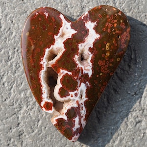 Puede incluir: Una piedra en forma de corazón con una superficie pulida. La piedra presenta una mezcla de patrones marrón rojizo oscuro y blanco, con pequeños puntos rojos circulares. El centro tiene una cavidad con formaciones de cristal blanco. La piedra mide unos 10 cm de largo.