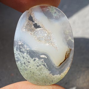 May include: Oval-shaped gemstone with a translucent appearance. The stone features a mix of white, light green, and brown hues, with a unique pattern of inclusions and textures. The gemstone is held between two fingers.