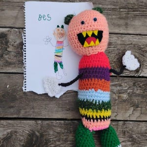 Puede incluir: Una muñeca de ganchillo hecha a mano con una cabeza rosa, brazos negros y un cuerpo a rayas en varios colores. La muñeca tiene una boca grande con dientes amarillos y está junto a un dibujo que dice "Bes".