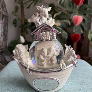 May include: A decorative snow globe featuring a boat-shaped base with animal figurines. The globe contains elephants and glitter, topped with giraffes and a house. The base is cream-colored with silver accents, creating a whimsical and charming aesthetic.