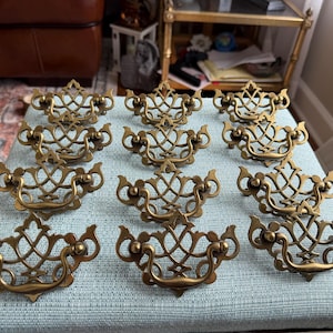 May include: A collection of twelve matching brass drawer pulls arranged in four rows of three. Each pull features an ornate, openwork metal design with a curved handle and a decorative backplate, showcasing a polished, aged metallic finish.