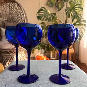 May include: Four cobalt blue wine glasses with rounded bowls and long stems are arranged on a light blue surface. The glasses are made of a reflective material, and the background includes plants and a framed sign.