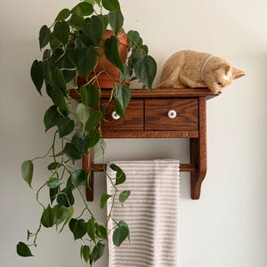 May include: A wall-mounted wooden shelf featuring two small drawers with white round knobs, a lower towel bar holding a beige and white striped towel, a potted heart-leaf philodendron plant, and a small tan cat figurine resting on top.