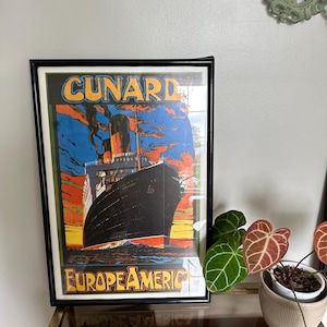 May include: Framed vintage travel poster with the text "CUNARD EUROPE AMERICA" featuring a large black ship sailing on blue water with a colorful sunset background. The poster is in a black frame.